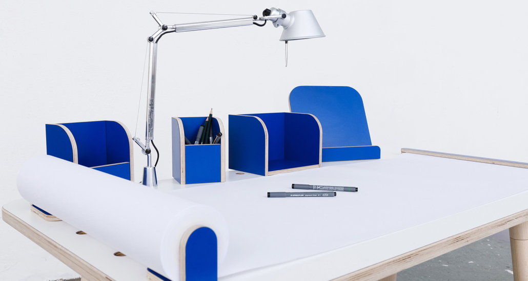 Ansicht auf den rowing table mit weißer Oberfläche und blauen Tools mit einer angebrachten Tischlampe sowie ausgerollten Papierrolle mit Stifen