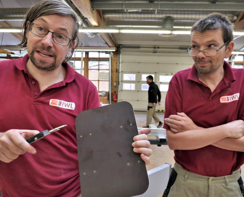 Zwei Mitarbeiter von pure position stehen in der Werkstatt und einer von den spricht in die Kamera mit einem Brett in der Hand